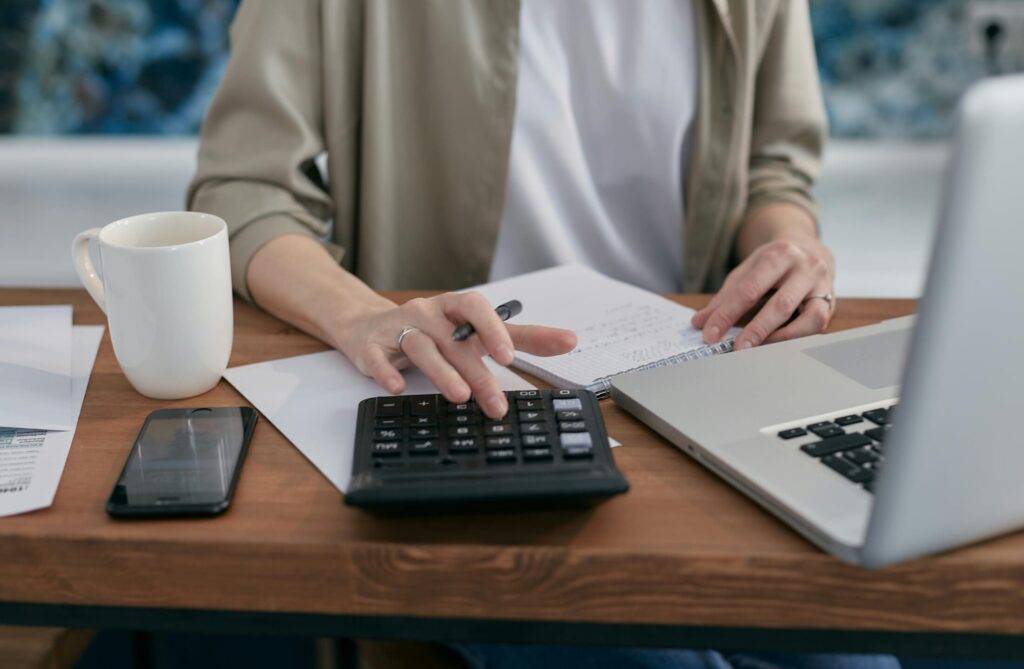a person sitting next to a wooden table with a notebook and laptop while using a calculator